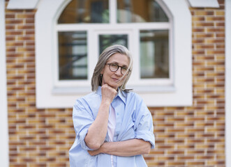 A woman in a blue shirt is standing in front of a brick building. She is wearing glasses and has her arms crossed