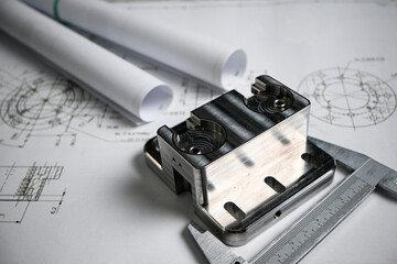 Metal machined part being measured with a caliper, placed on top of engineering blueprints