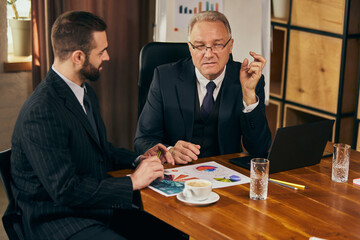 Senior businessman explaining data points to colleague, with charts and laptop on the table during team meeting. Personal professional consultation. Concept of business, teamwork, success