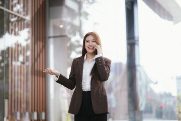 A casually dressed Asian businesswoman sips coffee while playing with her mobile phone outdoors....