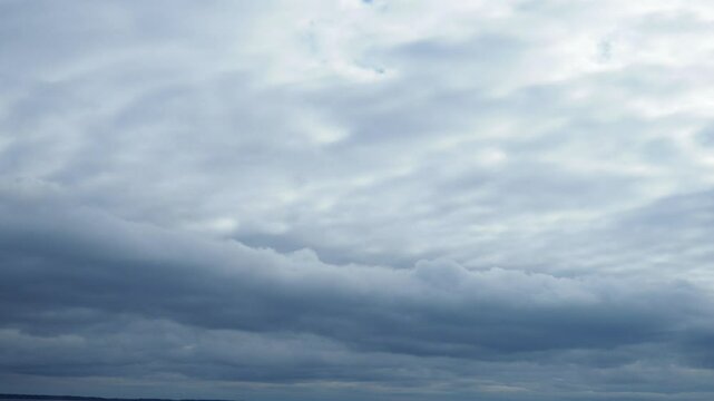 A low, long, heavy, oblong cloud floats across the sky above the lake. Lighter white and gray clouds fly above it. The two layers of clouds fly at different speeds and in different directions.
