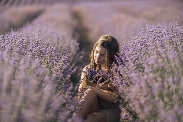 Girl is sitting in a field of purple flowers. She is holding a basket of flowers and smiling. Scene is peaceful and serene, as the girl is surrounded by the beauty of nature.