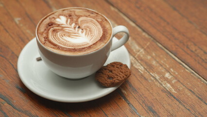 A cup of Cappucino coffee with a piece of chocolate biscuit on a wooden table.