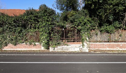 Rusty gate in the middle of a weathered  fence made of bricks and metal grid . Abandoned garden on behind,  road in front. Background for copy space.
