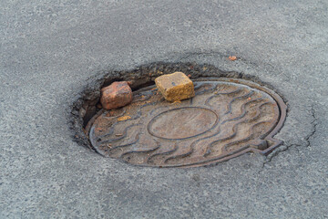 Street cast-iron metal manhole cover cap partially fell through the asphalt under excessive load. Two halves of red brick lie on it, next to it there are cracks from the deformation of the roadway