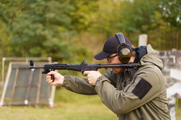 A man in a green jacket is holding a gun and wearing ear protection. He is in a grassy area with a few targets in the background