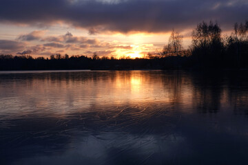 Beautiful natural landscape with thin ice near the shore, dramatic sunset and far forest trees silhouettes against calm river