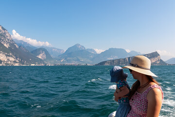 Mother with small toddler on ferry tour looking at lakeside town Torbole and Riva at Garda Lake, Trentino, North Italy. Surrounded by majestic mountain peaks of Garda Hills. Summer lakeside atmosphere