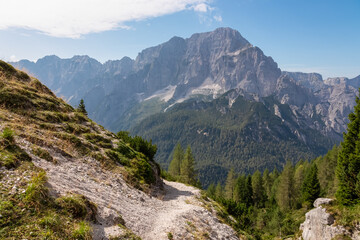 Fototapeta premium Hiking trail towards mountain peak Jof di Miezegnot with scenic view of rugged massif Jof di Montasio in Julian Alps, Friuli Venezia Giulia, Italy. Wanderlust in Italian Alps on sunny summer day. Hike