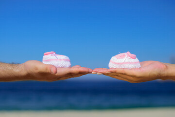 Hands holding pink baby shoes with a blurred beach and ocean background. Ideal for maternity announcements, family, and baby-themed concepts.