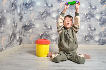 Naklejka premium A joyful boy in a tracksuit sits on the floor, holding a toy made of building blocks above his head