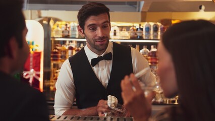 Focusing smiling smart bartender serving gin fizz or tonic soda at luxurious bar, engaging lively conversation with VIP customer couple at back view in atmosphere at lighting warm nightclub. Vinosity.
