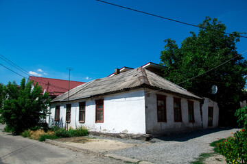 Old rural house with a weathered roof and wooden windows, surrounded by greenery and a clear blue...