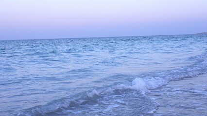 Fototapeta premium Ocean waves with soft white crests meet sandy shore under sky tinged with pre-dawn hues, creating tranquil scene evoking peace before sun rises. Morning calm concept.