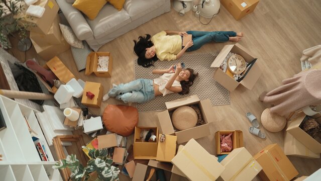 Lesbian lover looking at phone while laughing together and lying down at living room floor with cardboard box scattering. Happy couple checking at smart phone while spending time together. Myrmidon.