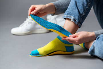 Close up of female hands holding blue orthopedic insoles, white sneakers on a gray background .