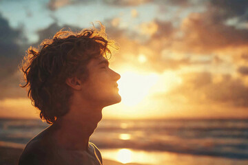 A young man with wavy hair enjoys the warm glow of the sunset on a beach his face turned upwards feeling the peaceful ocean breeze
