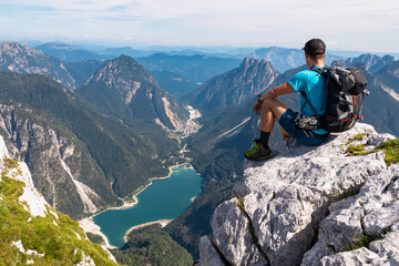 Naklejka premium Hiker man on top of Cima Del Lago with aerial view of alpine lake Lago Del Predil in Val Canale surrounded by majestic mountain peaks of Julian Alps, Friuli Venezia Giulia, Italy. Wanderlust in nature