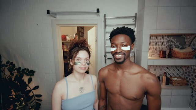 Young Caucasian girlfriend and African American boyfriend with under-eye patches standing in bathroom at home, posing on camera and smiling. Video portrait