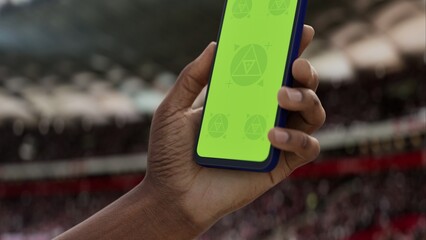 GREEN SCREEN CHROMA KEY A hand holding a phone with a green screen at a stadium during a football match. Ideal mockup for sports, betting, or event apps