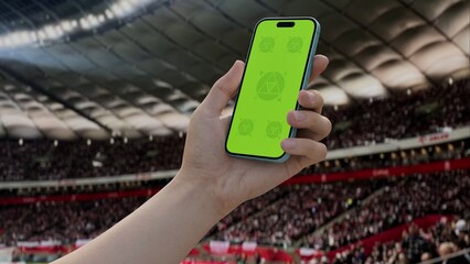 GREEN SCREEN CHROMA KEY A hand holding a phone with a green screen at a stadium during a football match. Ideal mockup for sports, betting, or event apps