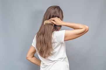 Fototapeta premium Wooden comb. Combing natural long hair. Rear view of woman in white t-shirt. Gray background, isolated.