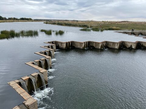 Calm river with gentle ripples overflowing from a low artificial dam with winding geometric shape, with visible bushes emerging from the water and the opposite riverbank, under a sky covered with thic