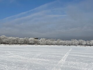 Winter scenery with frost-covered trees, partially cloudy sky, melting snow, a frozen lake in the...