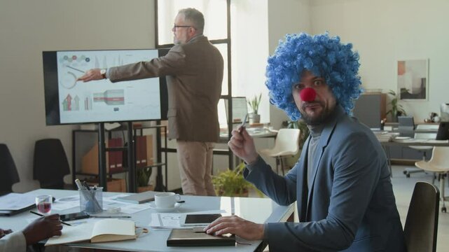 Funny businessman in clown wig and nose making silly faces on camera while CEO giving presentation at meeting in office