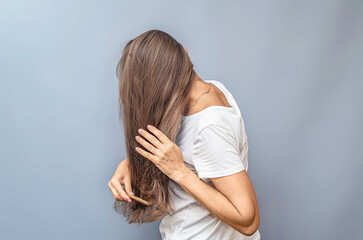 Fototapeta premium Side view of combing long hair. Woman in white t-shirt. Gray background, isolated. Hair care problem.
