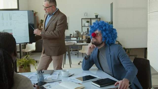 Hilarious businessman wearing clown wig and nose listening to presentation of colleague, nodding his head, then crossing arms and expressing disagreement during team meeting in office