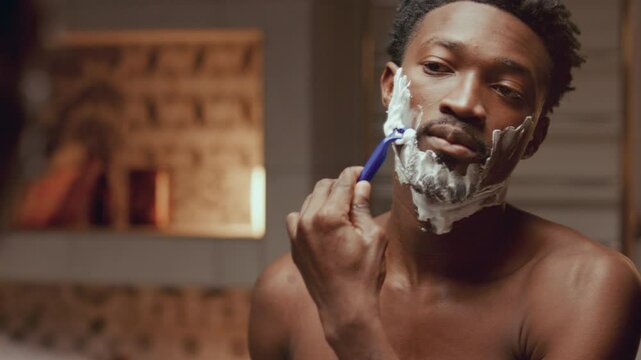 Young Black man with shirtless torso standing in bathroom at home, looking in mirror and shaving his face with razor during daily routine