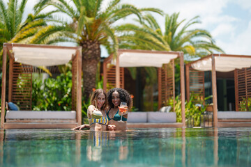 Two joyful friends, one mixed race and one Caucasian, enjoy sunny leisure time in a pool, smiling...