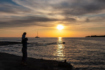 Loving mother with toddler at coastline of Adriatic Mediterranean sea during sunset. Luxury sailboat silhouetted against fiery sky. Calm Bay of Dajla in Karigador, Istria peninsula, Croatia, Europe