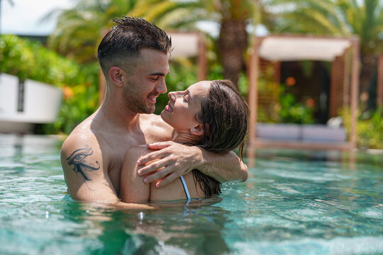 A cheerful young Caucasian couple embrace warmly in a sunny pool, the man's tattoo visible as they share a tender, loving interaction, enhancing the serene outdoor setting.