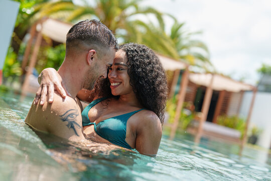 A cheerful man and woman, diverse in ethnicity, share a loving embrace in a sunlit pool, their swimwear hinting at leisurely summer activities in a tropical location.