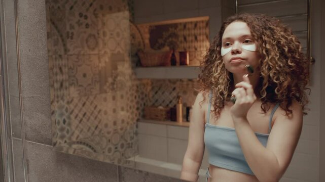 Young curly-haired girl with under-eye patches, standing in front of bathroom mirror and doing facial massage with jade roller during beauty routine