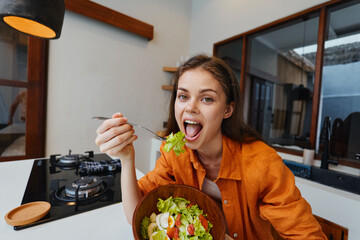 Young woman enjoying a healthy salad in a cozy kitchen, wearing an orange shirt, expressing joy and...