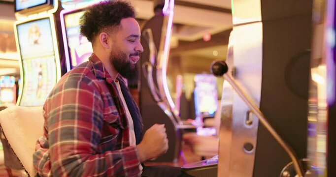 A young man is overjoyed after winning at a busy casino, celebrating a significant slot machine victory. The vibrant atmosphere is filled with energy from other players and friends around him