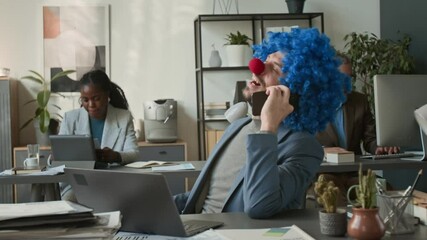 Goofy businessman in clown wig and nose answering phone call from unpleasant client, pretending to speak politely to him at desk in open-plan office