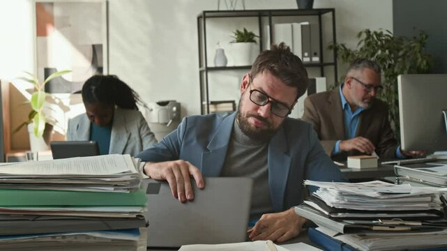 Overloaded office worker putting on clown wig and nose, then typing on laptop with fake smile at desk with huge piles of papers stacked on top of it