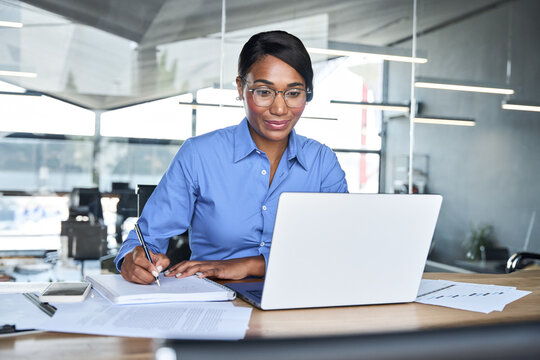 Busy professional business woman ceo executive working on computer in office. Young African American female manager or employee using laptop writing notes sitting at desk elearning online. - Powered by Adobe