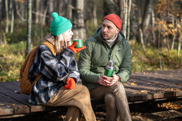 Fototapeta premium Satisfied best friends, hiker travelers sitting on wooden eco path trail through September thicket, talking, chatting, speaking, discussing various topics with interest in pleasant atmosphere.