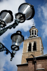 Low-angle view of a historic church tower, with ornate street lamps in the foreground.  Sunny day, blue sky, and clouds.