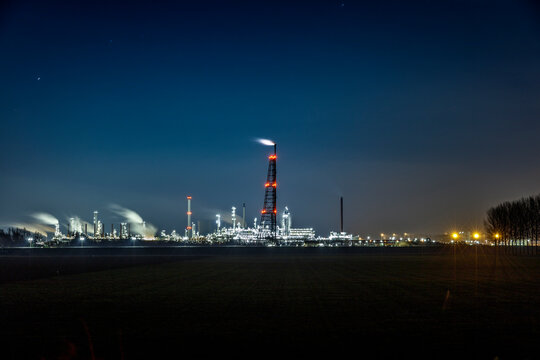 Nighttime view of a large industrial complex, illuminated by lights, with smoke stacks emitting vapor.  Industrial landscape at night.