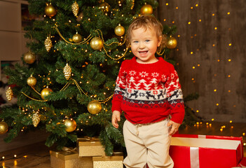 Christmas Child next to Christmas Tree decorated with Golden Ornament in Cozy Interior. Happy smiling Baby Boy in red knitted Xmas sweater in Room with Gifts