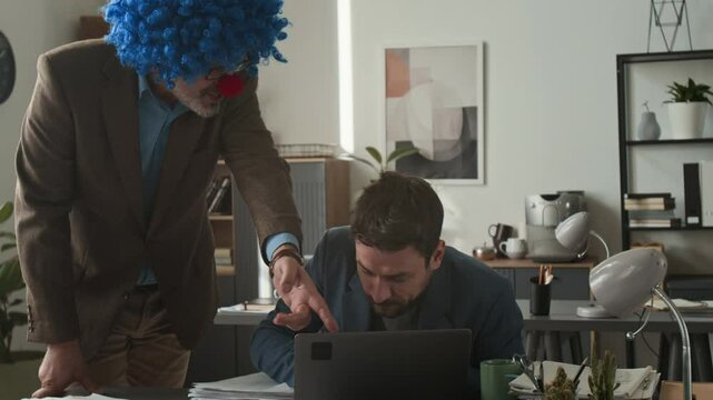 Annoyed boss dressed like clown shouting at scared employee, throwing pile of papers on his desk and giving strict instructions during workday in office