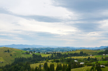Fototapeta premium Beautiful panoramic view showcasing rural houses and rolling green hills against a backdrop of a cloudy sky. Ideal for depicting tranquility, nature, and countryside landscapes. Carpathians, Ukraine 