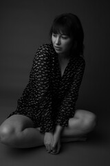 Black and white studio portrait of a caucasian woman with long dark hair with bangs. She is sitting on the floor cross legged wearing a long sleeve pleated dress. She is looking at the camera.