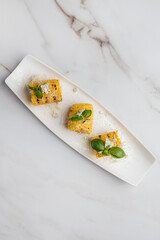 Three pieces of grilled corn on the cob are served on a white rectangular plate, topped with blue cheese, basil leaves, and grated cheese. Restaurant or cafe menu, cookbook recipe


